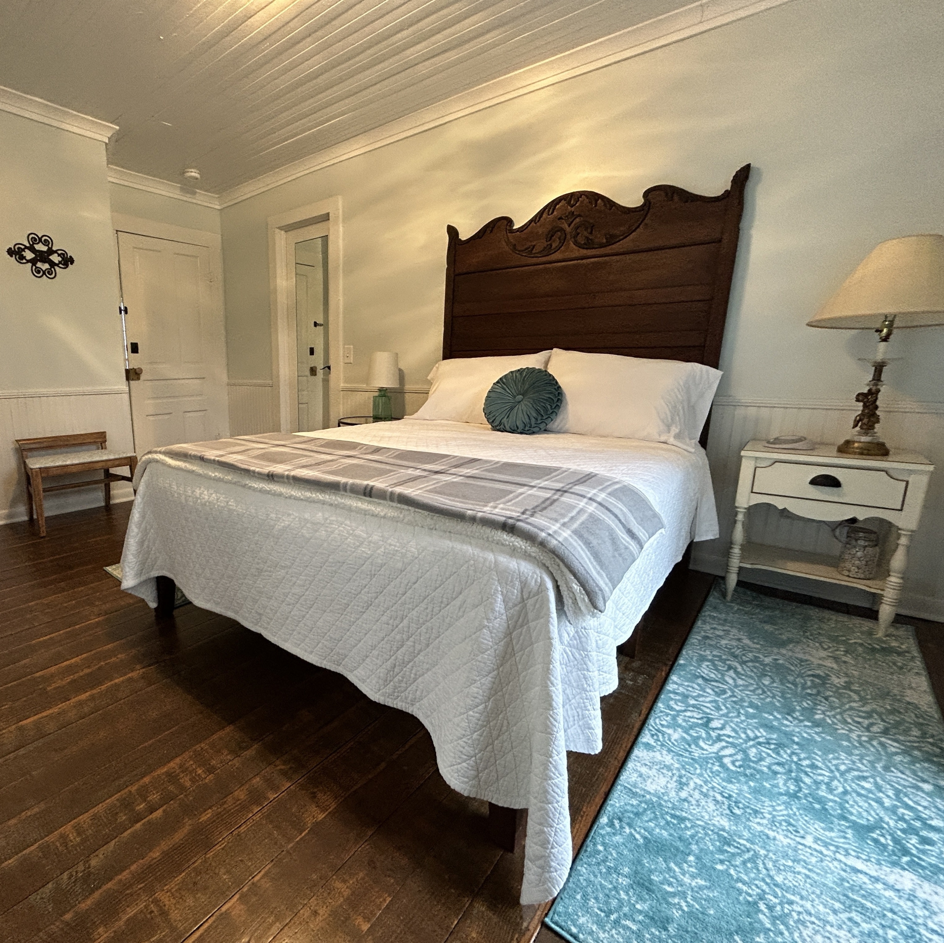 Bed with wooden headboard, white bedspread with grey blanket and round teal pillow. Table with lamp on the right side.