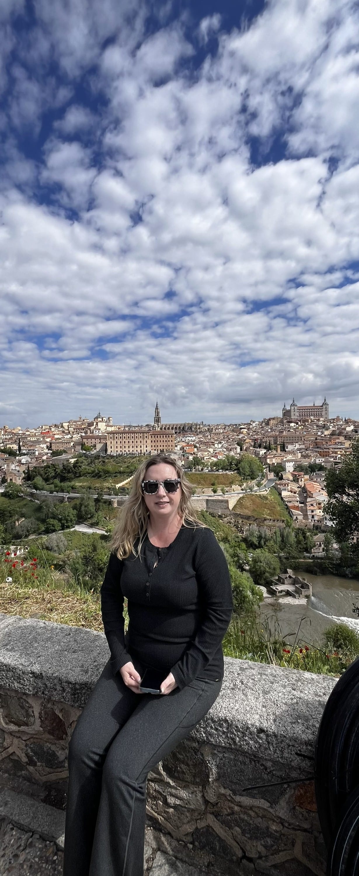Stephanie, wearing sunglasses and a black top sits on a stone wall overlooking the historic city of Toledo, Spain, with drama