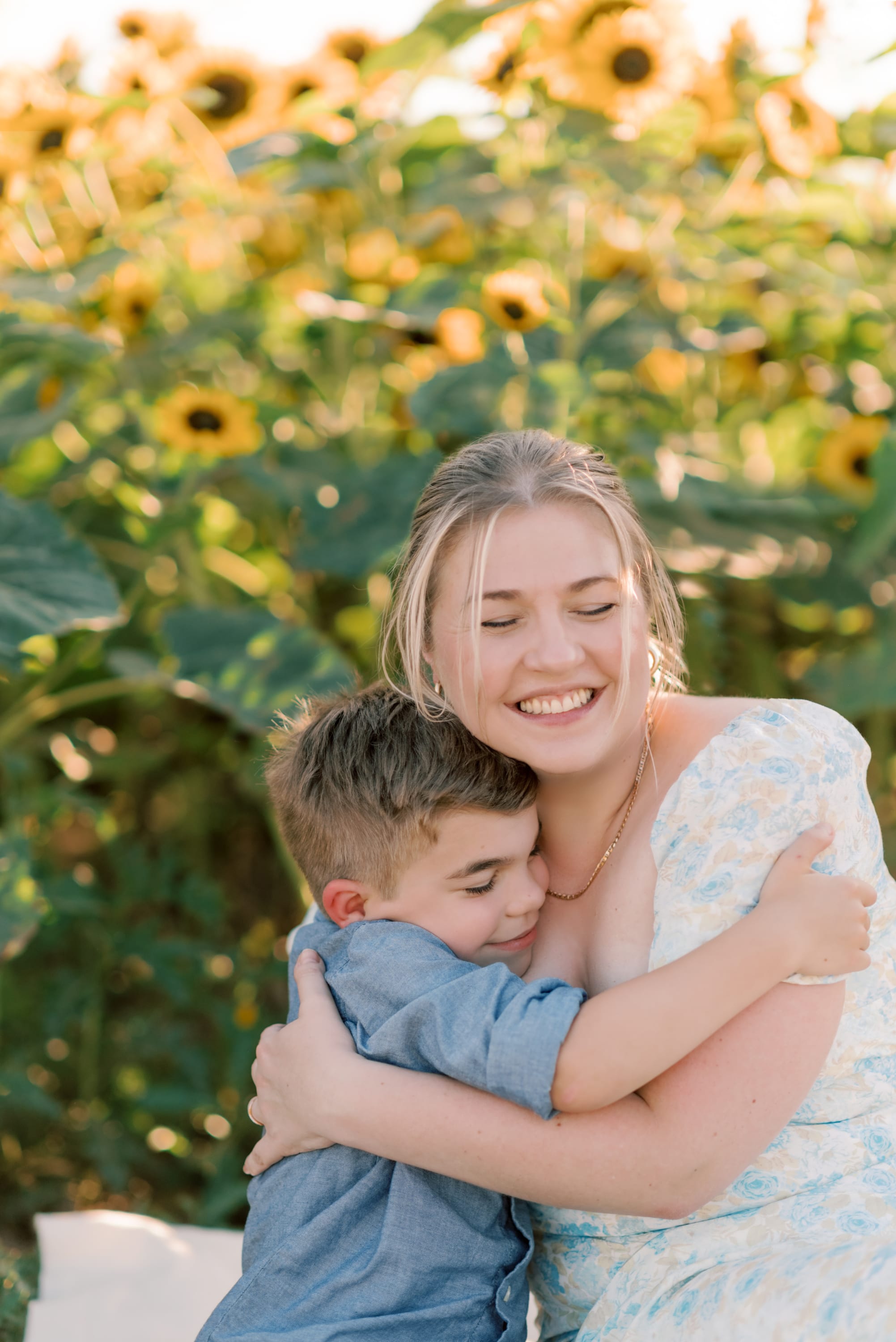 Mom in white floral dress hugging her young son in a field of sunflowers