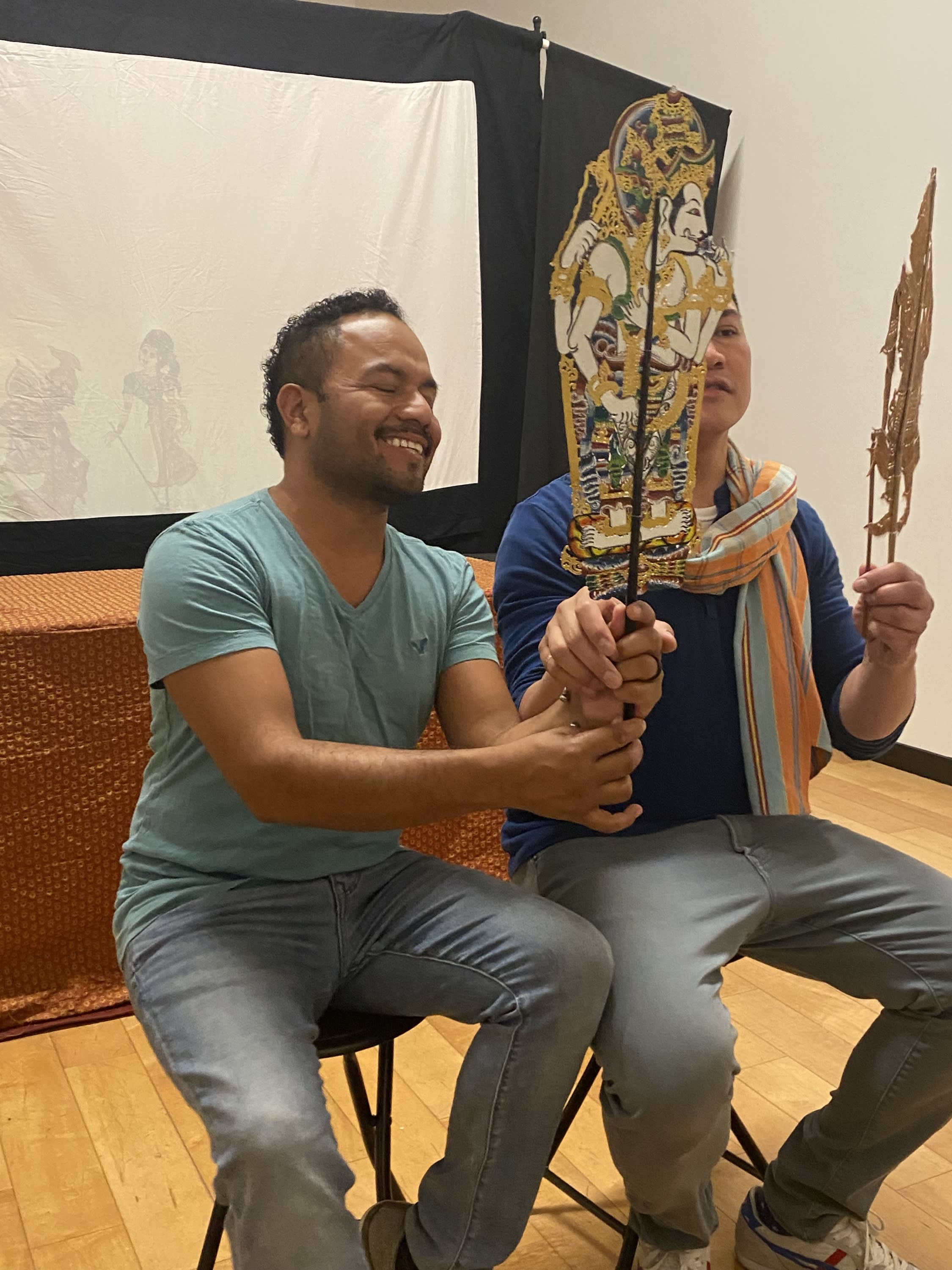 2 male actors sit on stools holding traditional Thai shadow puppets