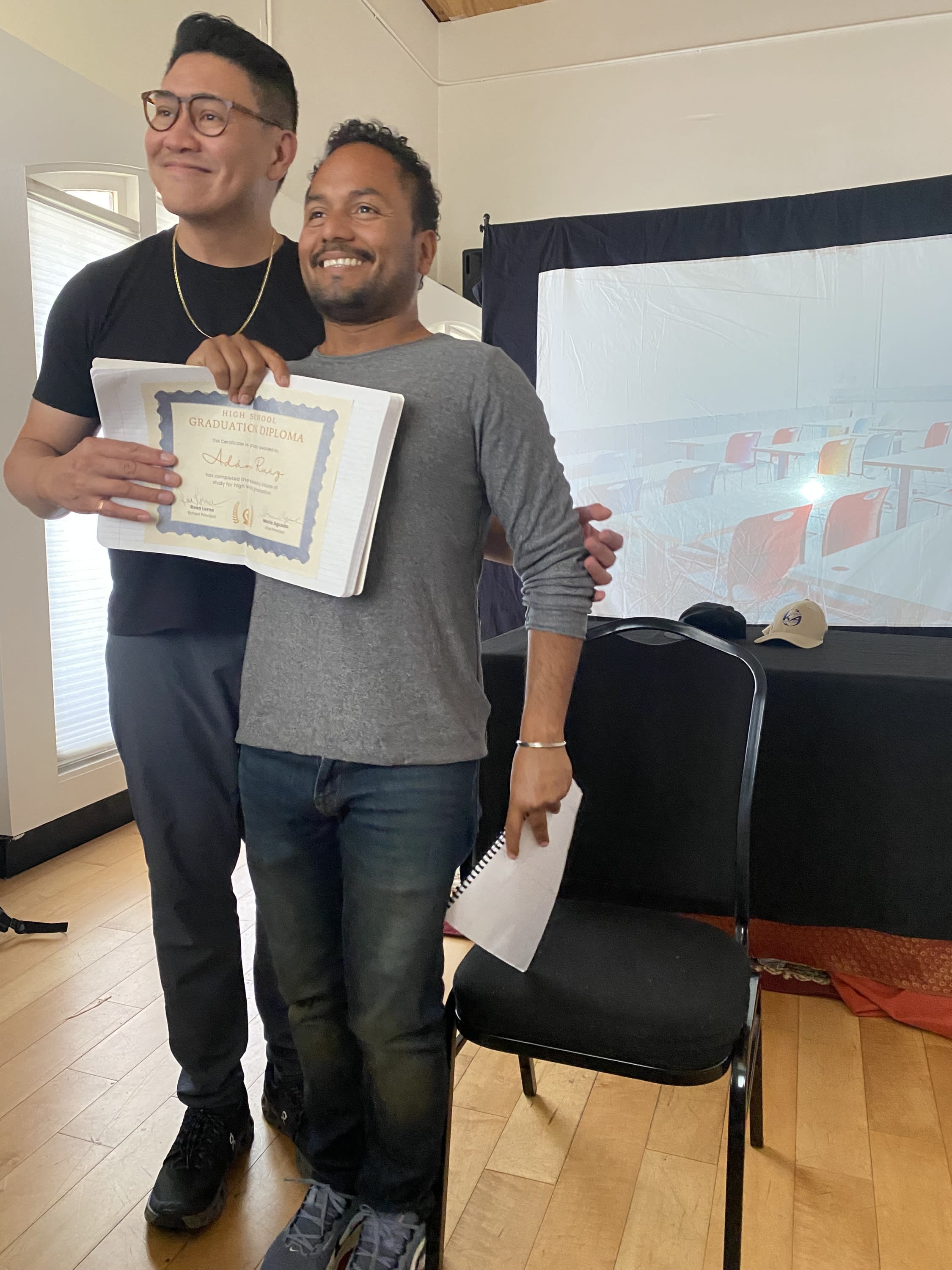 Two actors stand smiling and holding a diploma as if posing for a picture.