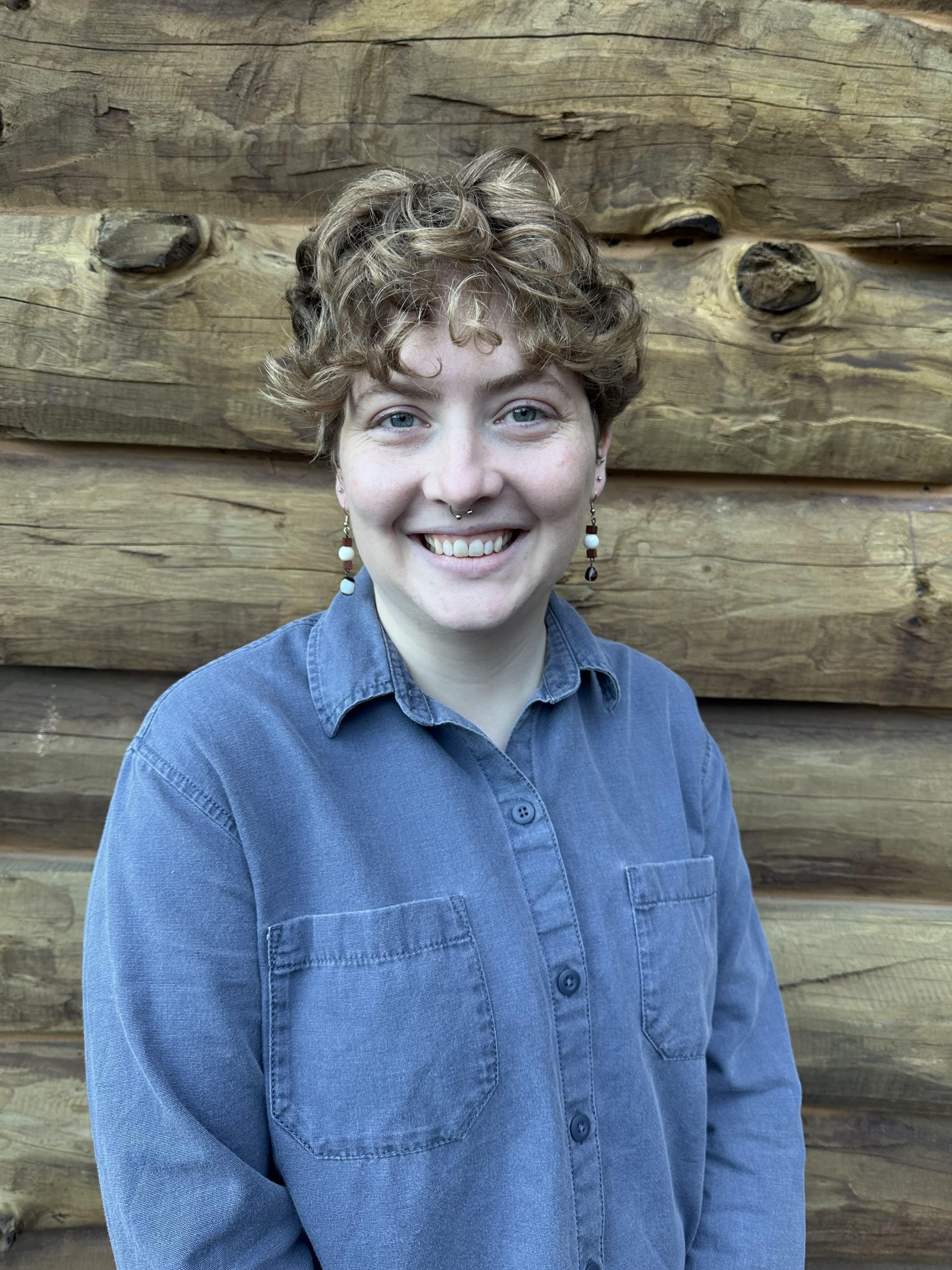 A white person smiling with blonde/brown curly hair wearing a blue collared long sleeve shirt.