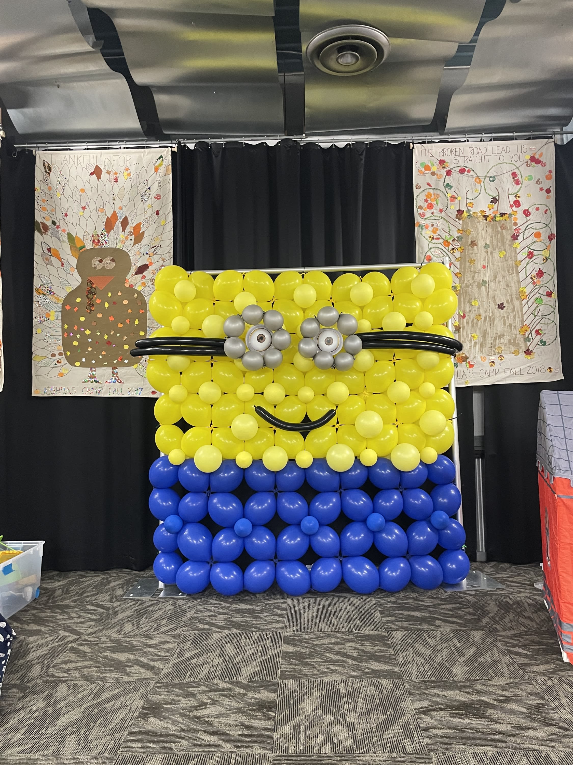 Balloon backdrop made to look like a Minion character, using yellow and blue balloons with silver balloon eyes and a black sm