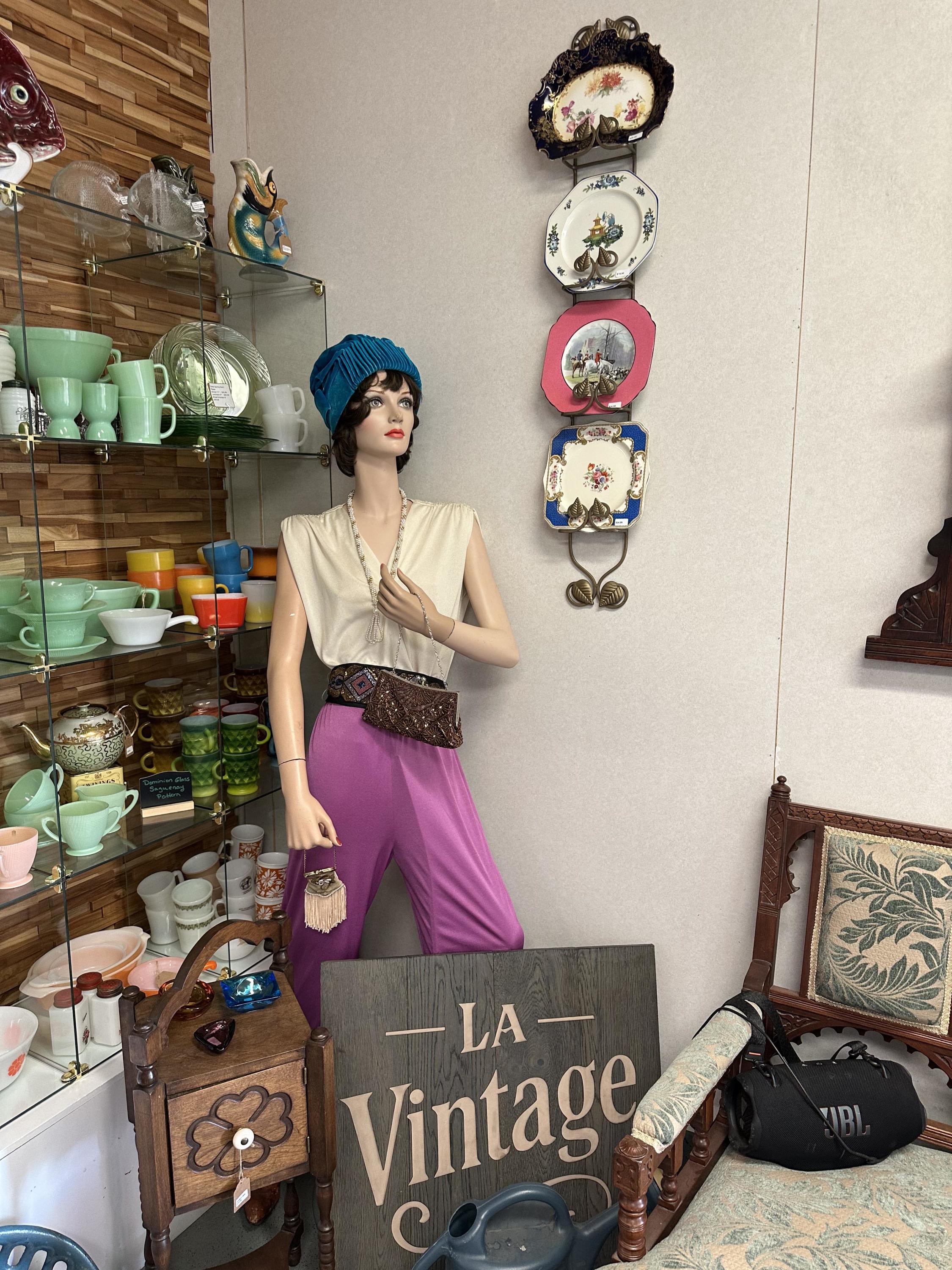 Mannequin in retro outfit surrounded by vintage glassware, collectible plates, and antique furniture inside LA Vintage.