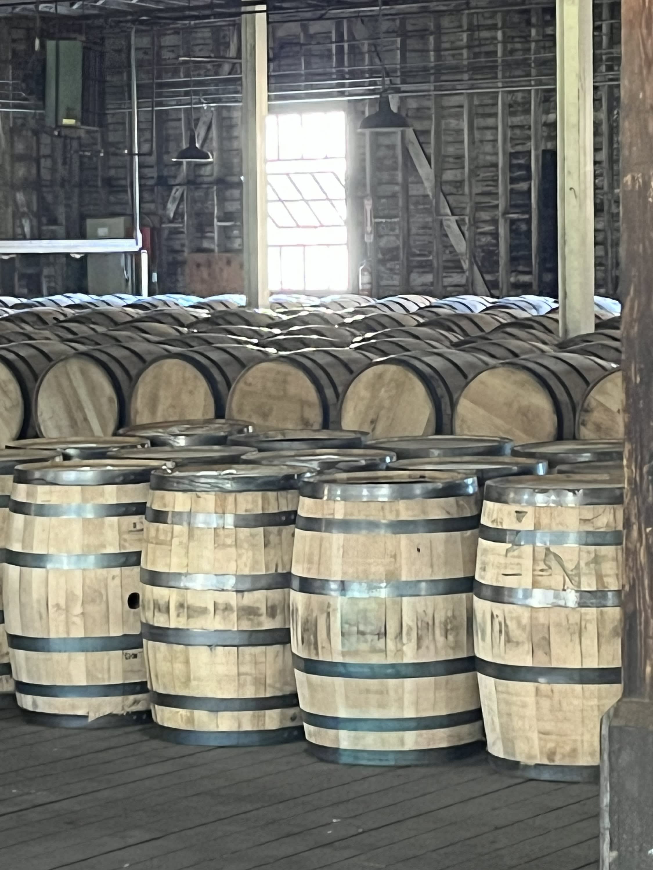 Rows of wooden bourbon barrels inside a rickhouse, stacked and aging in a dim, rustic warehouse.