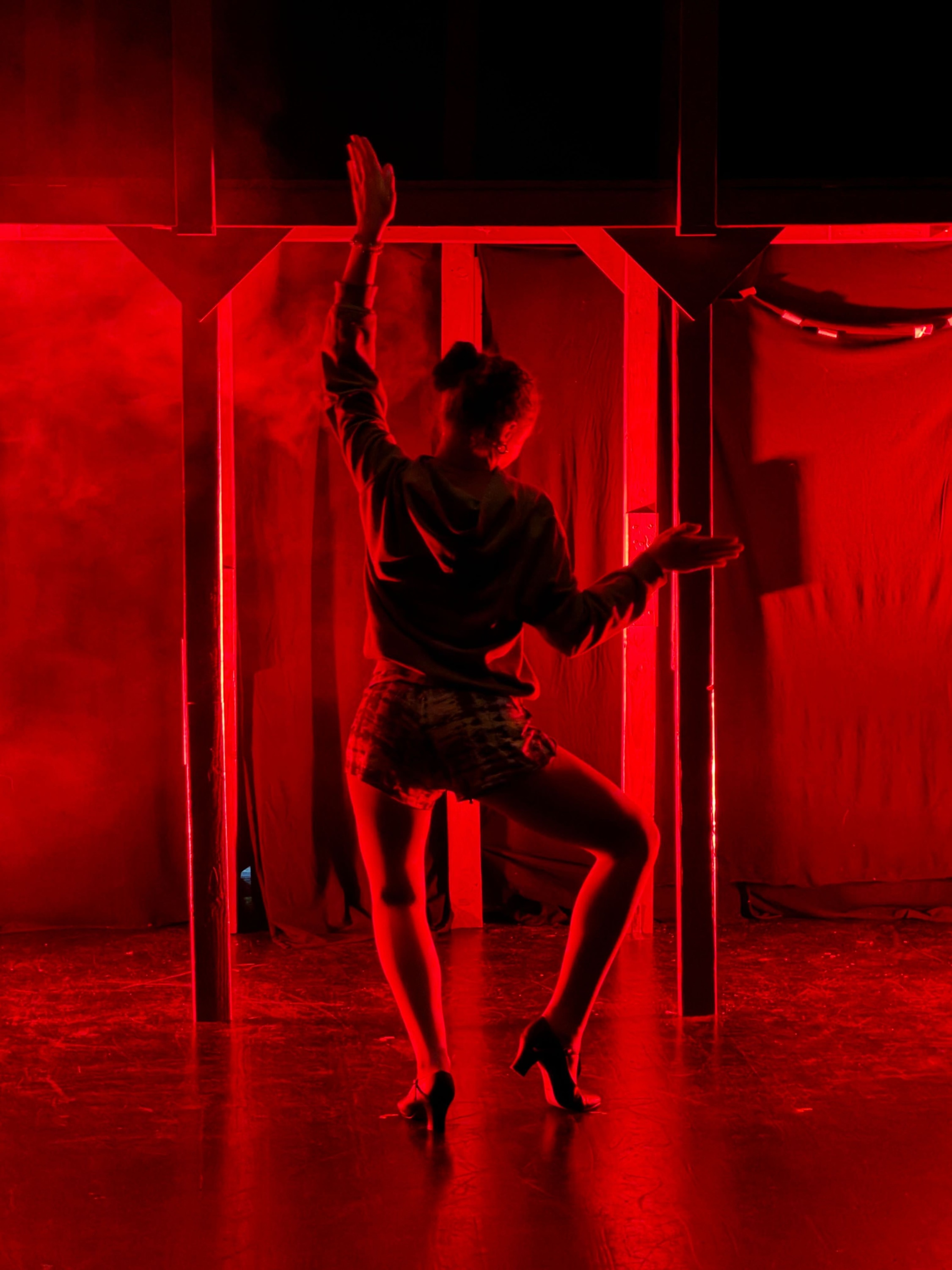 Dancer posed with Fosse-style hands up in the air in front of a bleacher-type structure, with fog and red/black lighting
