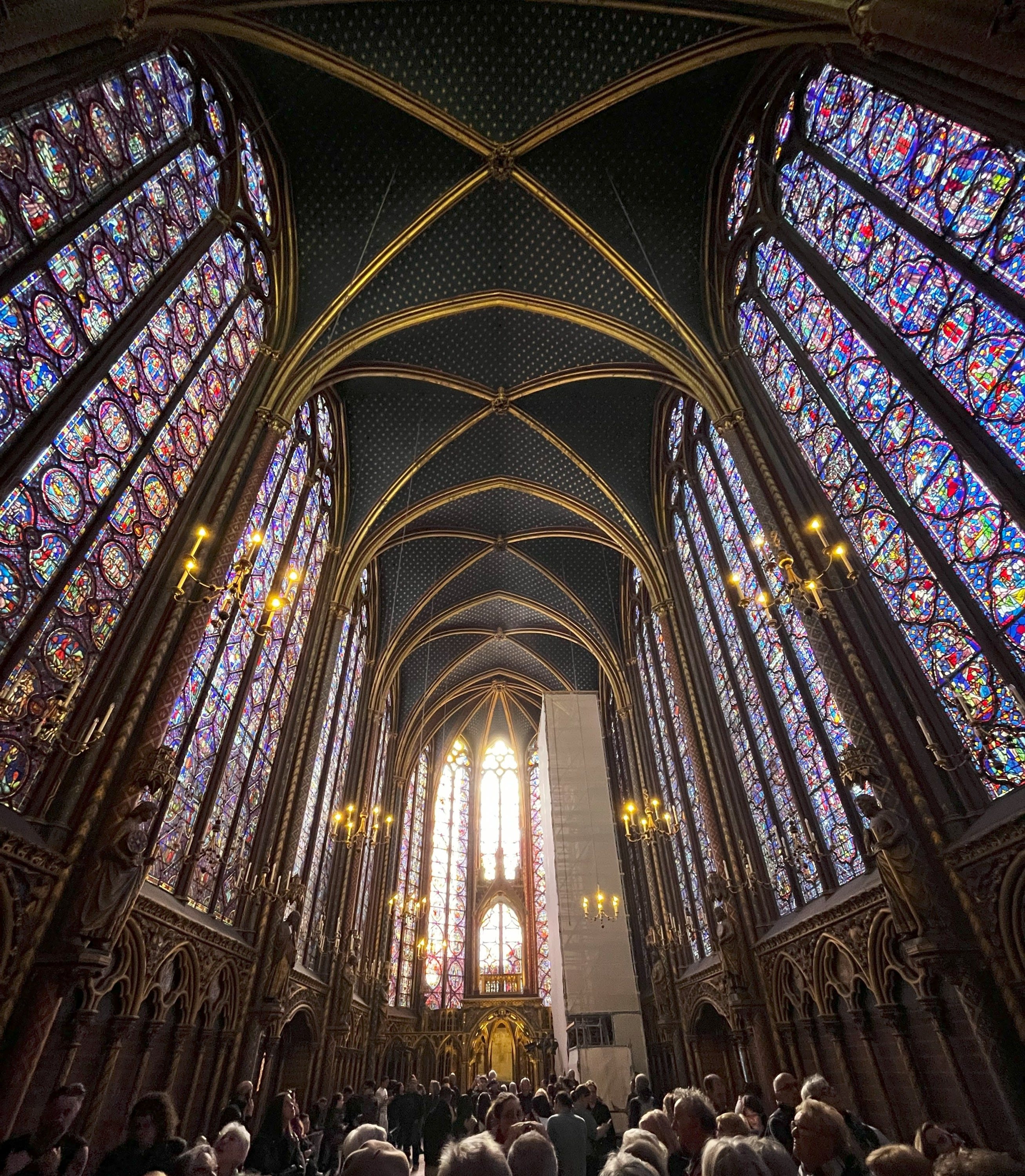 Interior view of Sainte-Chapelle in Paris, showing tall stained-glass windows, vaulted ceilings, chandeliers, and a crowd of