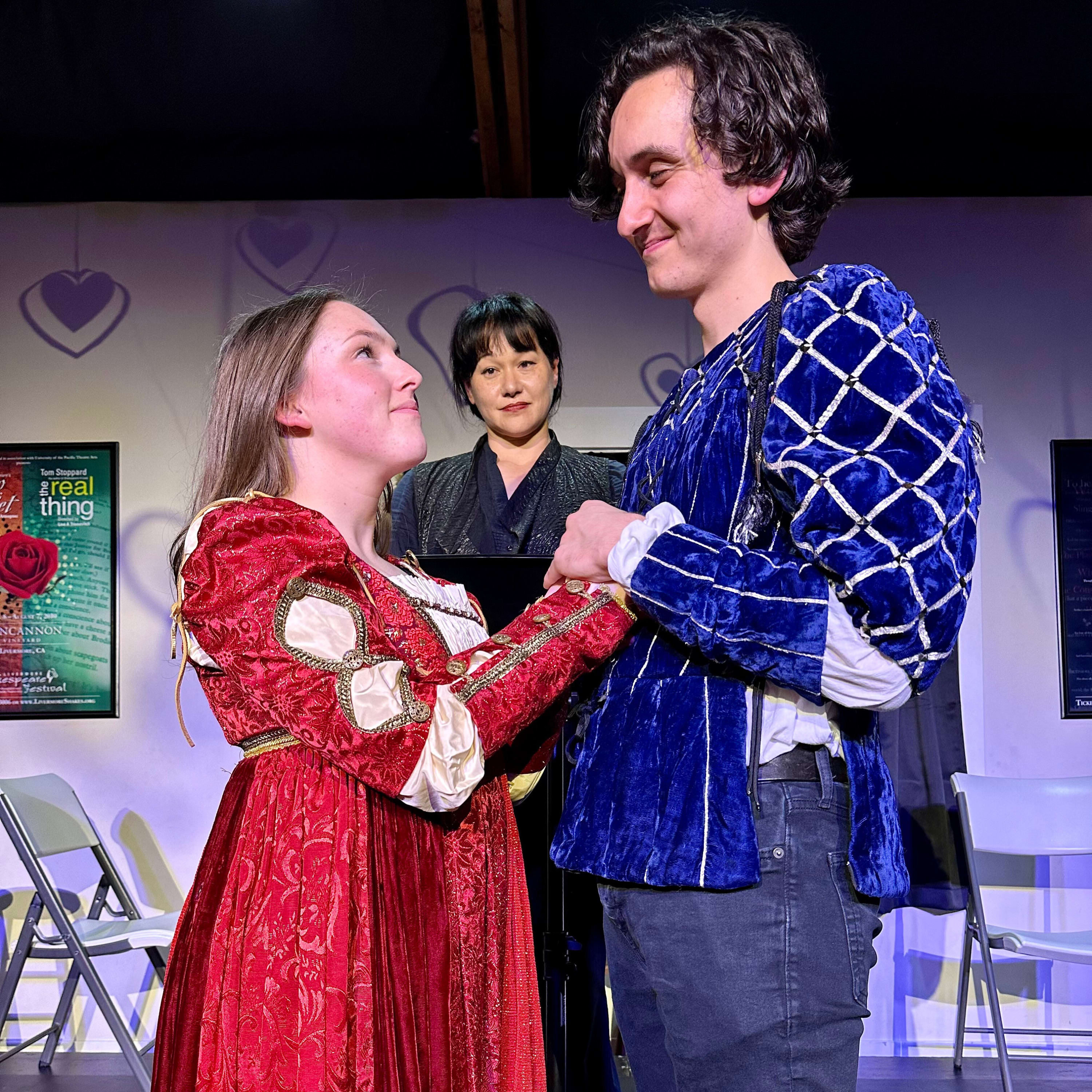 A woman (Juliet) and man (Romeo) in 14th century style costumes hold hands and gaze at each other lovingly. A woman stands be