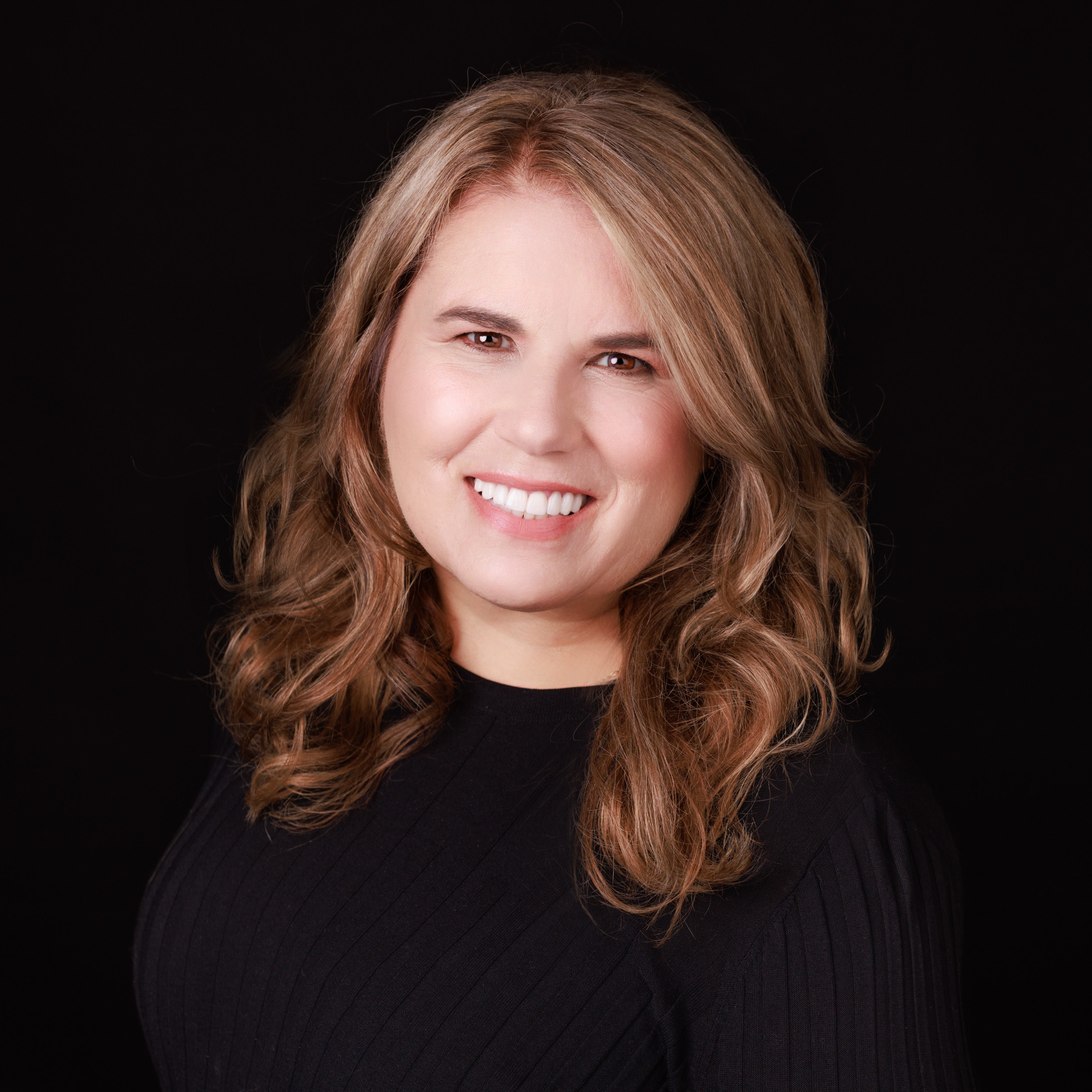 Anne Sheldon headshot, blonde hair, brown eyes, smiling, wearing black sweater