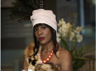 A young woman wears a headcovering studded with peacock feathers, and wears a pink faux-pearl necklace