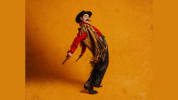 a woman dressed as a male cowboy throws his fringe back against an orange background