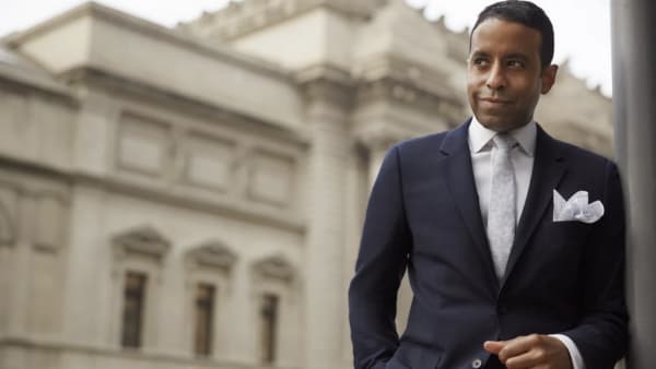 A gentleman in a suit and tie standing on the right corner of the image, smiling as he looks towards the left. A blurry build