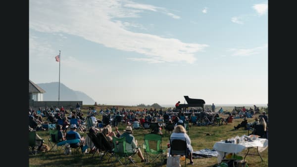 Hunter Noack sits at a piano surrounded by audience who members are seated in camp chairs watching Hunter play.  There is a f