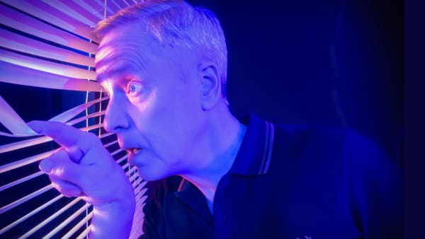 John Fisher peeks out of venetian blinds, with blue and purple light tinting the image