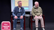 Two men sit in chairs representing a car, with a projection screen behind them. One wears all blue, and the other wears a gre