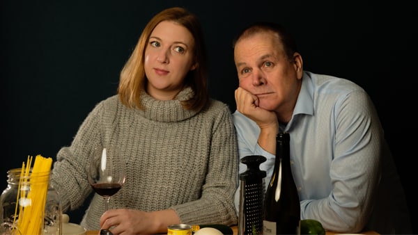 A white woman with short brown hair, a grey sweater,  and a wine glass next to an older white man with a light blue button up