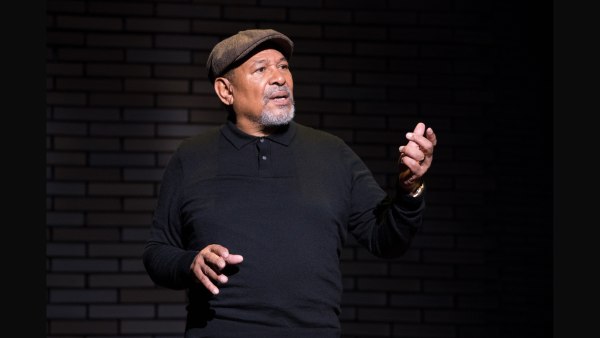 An actor in black shirt and brown cap gestures toward the audience in front of a brick wall