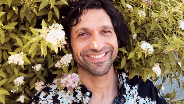 Man with light skin and dark hair smiles leaning against a wall of greenery and flowers