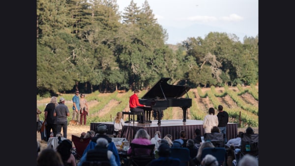 Hunter Noack plays piano surrounded by audience members seated in camp chairs, on the ground or walking around