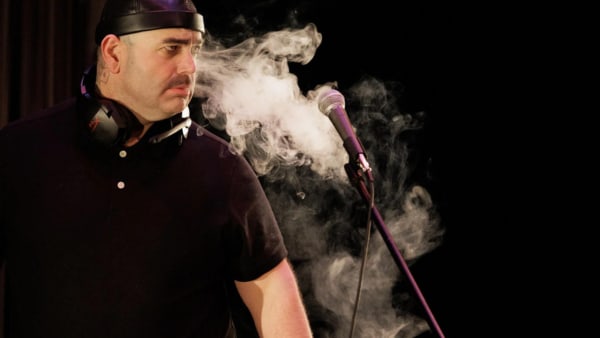 A large person dressed in black has smoke billowing around them, coming out of their face towards a microphone in front of th