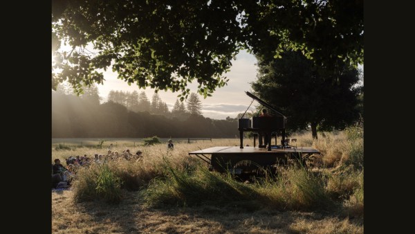 Hunter Noack sits at a piano at sunset in a lush green area surrounded by trees. Audience members are seated on the ground w