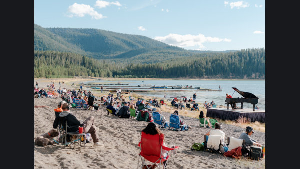 People in camping chairs surround Hunter Noack playing the piano on a beach.  There's boats, a lake, and tree toped