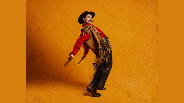 a woman dressed as a male cowboy throws his fringe back against an orange background