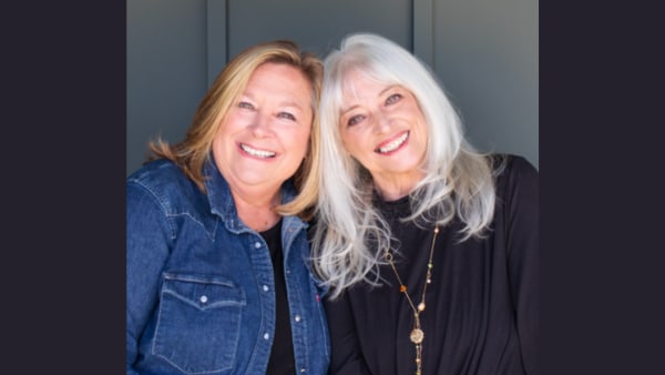 Two women with heads leaned together. One has blonde hair and a blue jean jacket. The other has white hair and a black shirt.