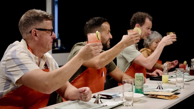 A group of people sitting at a table, each holding up colorful cocktails in a toast. They are wearing orange aprons.