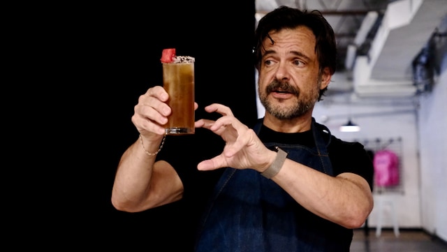 A man with a beard and apron holds up a tall cocktail glass filled with a dark beverage and garnished with a slice of fruit.