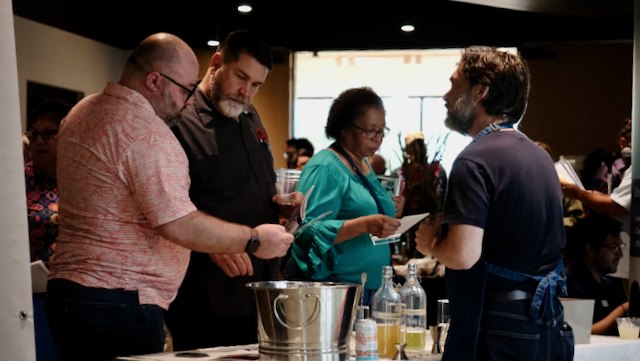 A group of people gathers around a table in a warmly lit space, conversing and engaging over documents and beverages.