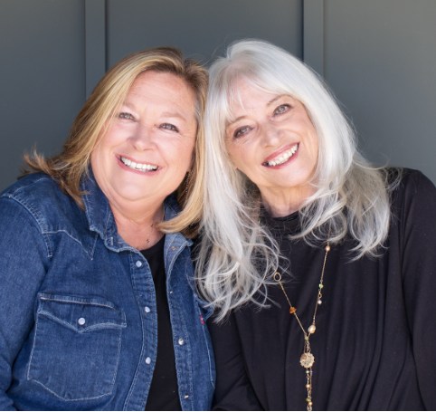 Two women with heads leaned together. One has blonde hair and a blue jean jacket. The other has white hair and a black shirt.