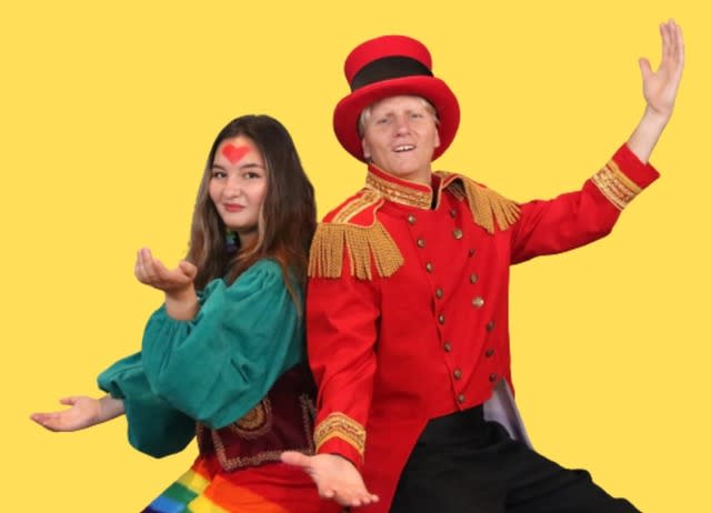 an actor in a red army jacket and an actor in an aqua bouse pose together in front of a yellow background