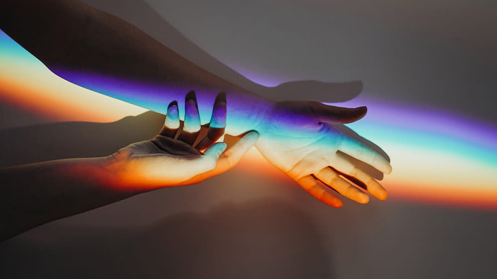 Rainbow colors are projected onto two arms against a gray background. One hand reaches toward the other arm with fingertips l