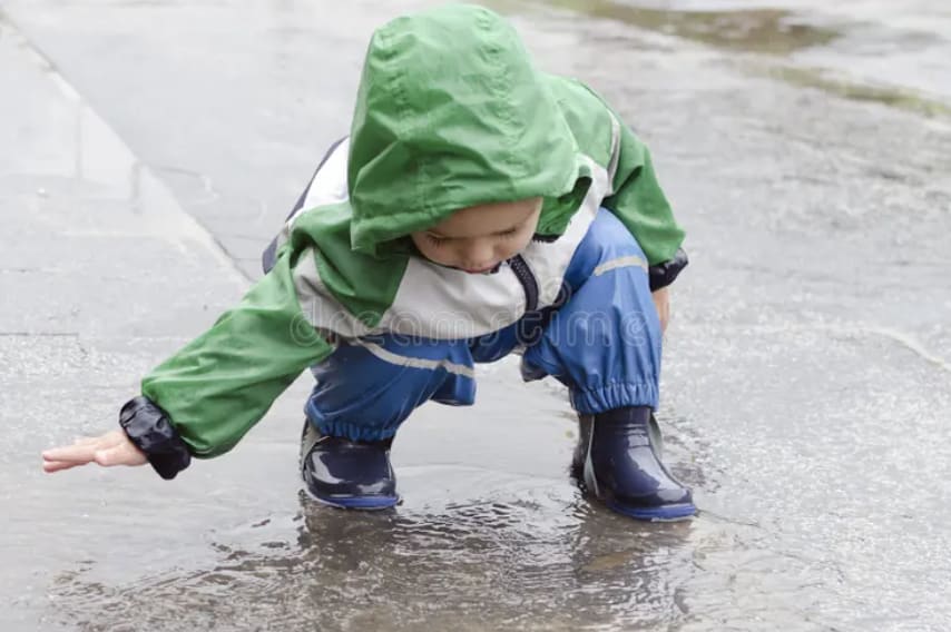 Child splashing in puddle with green rain jacket and blue runner boots