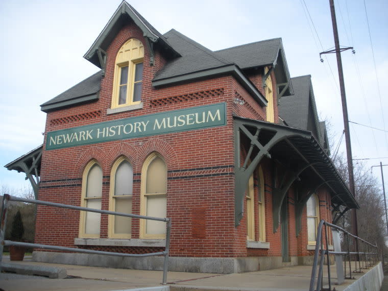A red brick building with a gray roof. A green sign with white lettering says Newark History Museum