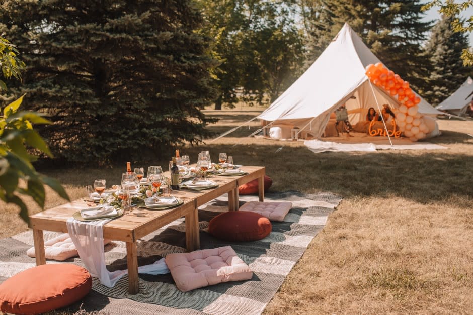 Outdoor bridal picnic setup with a low wooden table, wine glasses, and floor cushions next to a tent decorated with orange ba