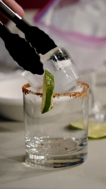 Close-up of a hand using black tongs to place a large ice cube into a clear glass with a chili powder rim.