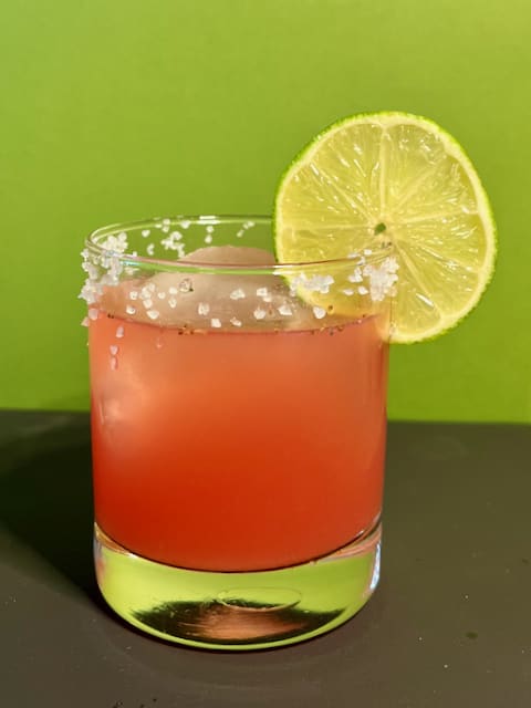 A vibrant pink cocktail in a clear glass with a salted rim, garnished with a fresh lime slice on the edge.