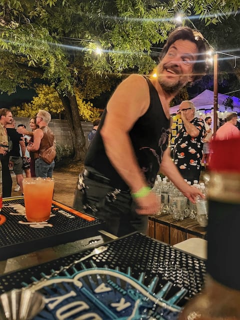 A lively bear-tender in a black tank top is caught mid-motion while serving drinks at an outdoor bar during the evening.