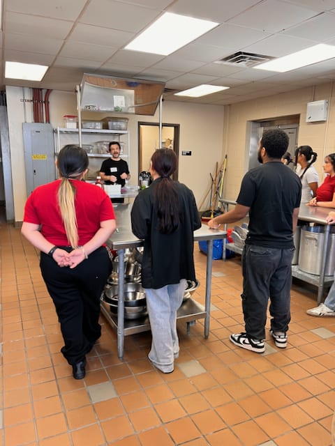 A group of people stand attentively around a table in a commercial kitchen, facing a person giving a presentation.
