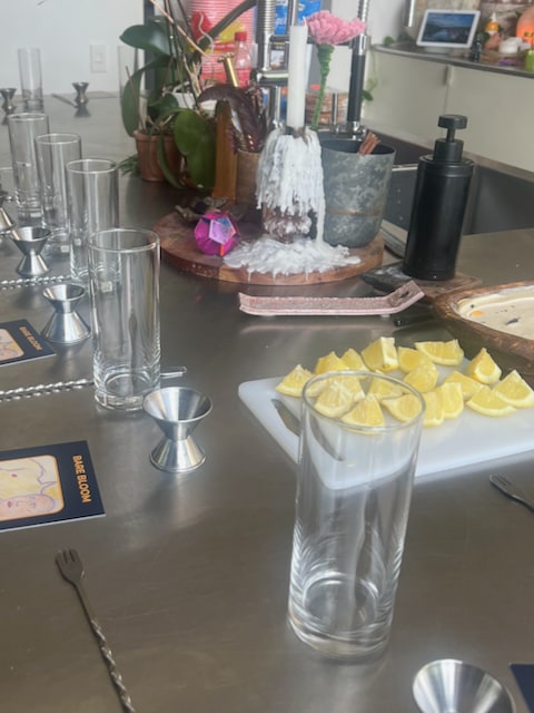 A kitchen countertop setup featuring tall glasses, cocktail jiggers, lemon wedges on a cutting board, & bar accessories.