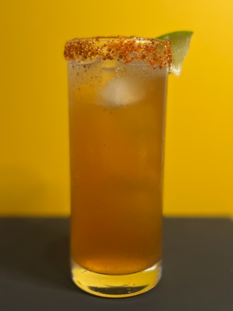 A tall glass of amber-colored ENAB garnished with a lime wedge and a rim coated with chili powder and seasoning.