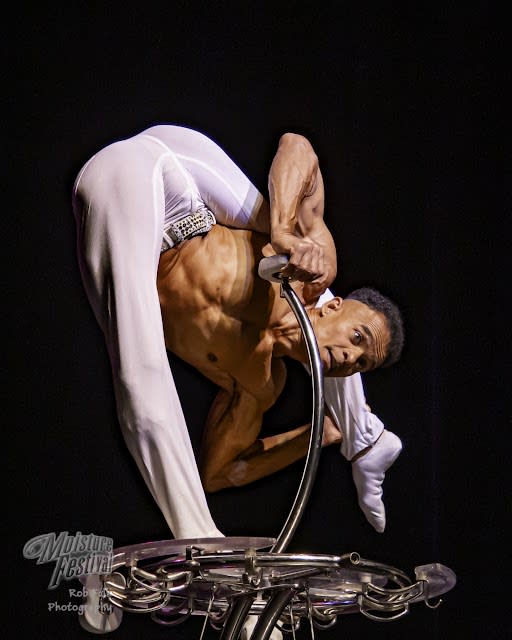 A bare-chested, light-skinned brown man wearing white pants is standing on one leg in a wild and indescribable contortion