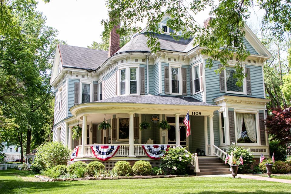Light blue two-story vintage mansion with white wraparound porch, American flag and bunting