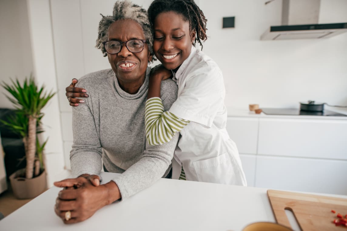 two women smiling in a kitchen setting, one hugging another