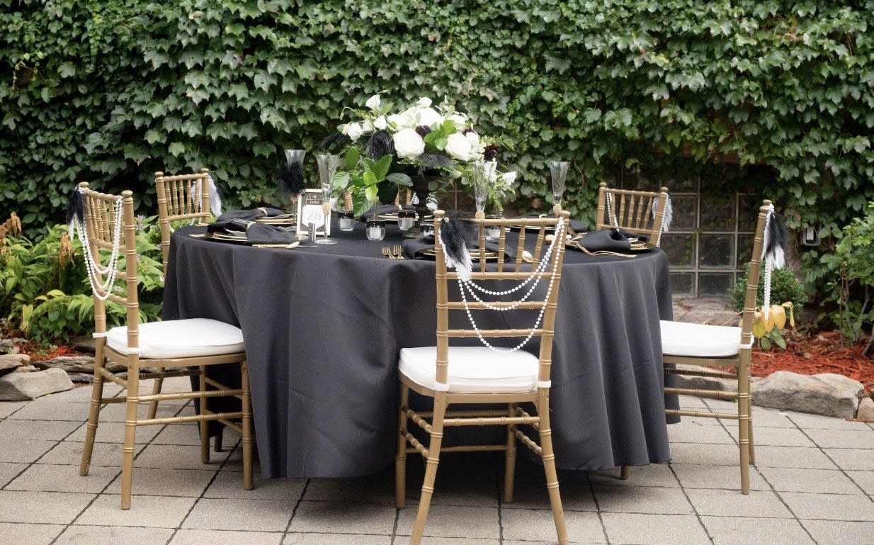 Outdoor table setup with black linens, white florals, and gold chairs adorned with pearls and feathers.