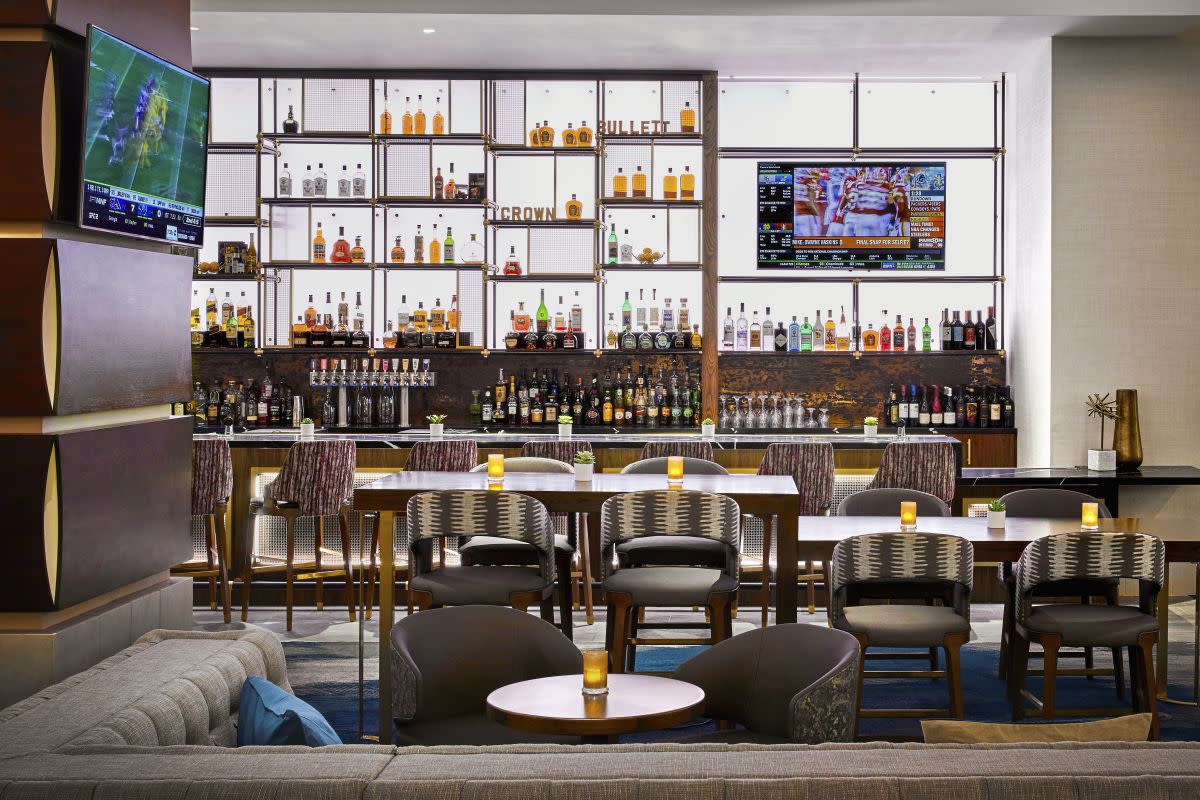 JW Marriott New Orleans Bar. Shows a backlit wall with alcohol bottles, a sleek silvery bar, and lux booths and club chairs.