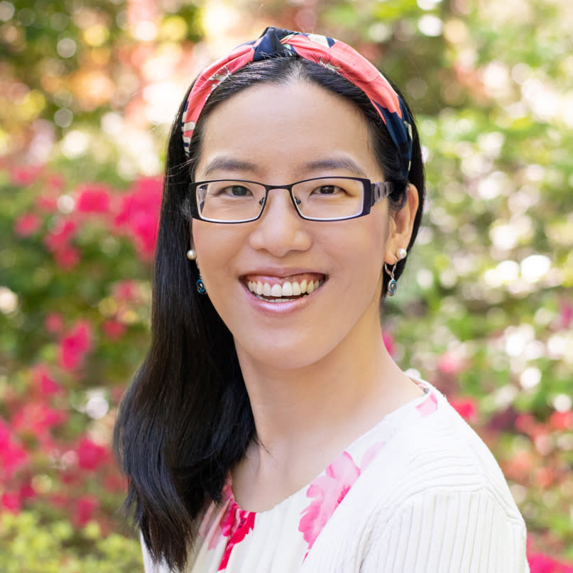 Asian women smiling headshot at a park with color flowers in the background.