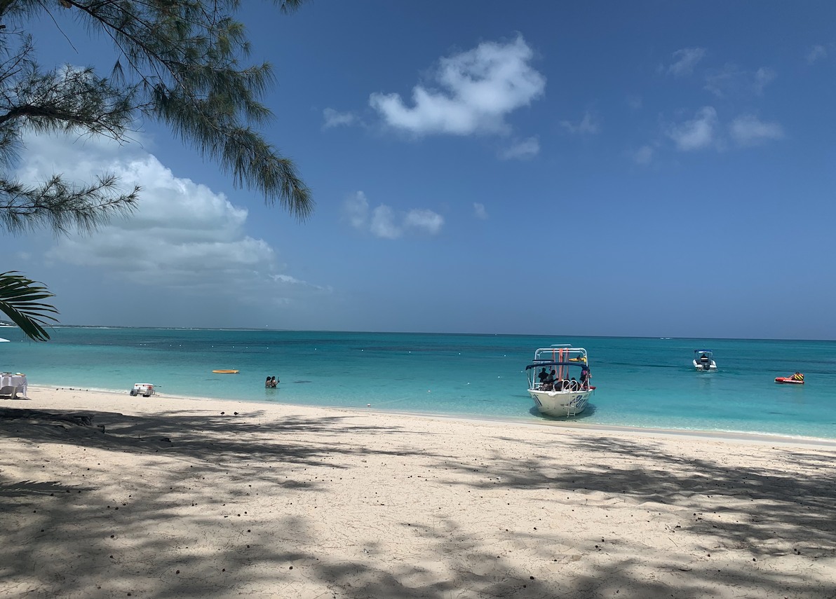 A white sand beach, next to the Caribbean sea.  There are a couple of boats and some swimmers in the water.